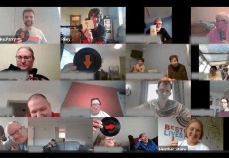 A video call grid showing multiple people in separate home settings. Several participants are holding up circular black signs or cards with orange arrows pointing up or down, while others hold paper cards or sit watching.