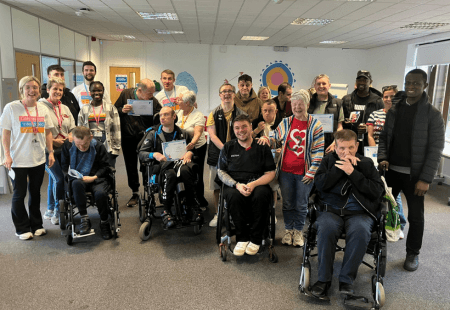 A large group of people gathered indoors for a group photo, several using wheelchairs, standing and sitting together in a bright community room with posters on the walls, with some holding certificates.