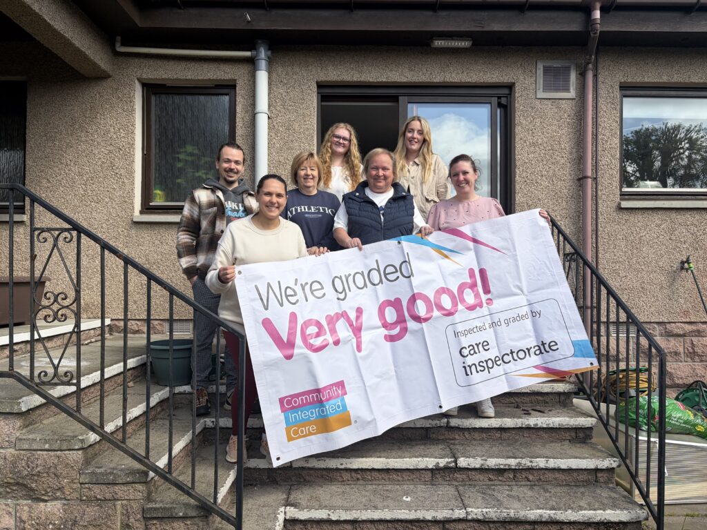Colleagues are smiling and holding up a banner reading 'We're rated Very Good by the Care Inspectorate'.