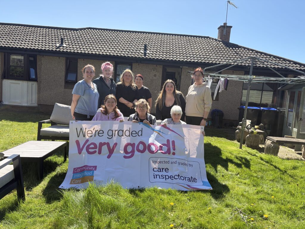 Colleagues are smiling and holding up a banner reading 'We're rated Very Good by the Care Inspectorate'.