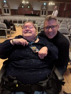 Judith, smiling in her wheelchair, next to TV presenter Ben Shephard at a venue, both looking happy as they smile facing forward at the camera.