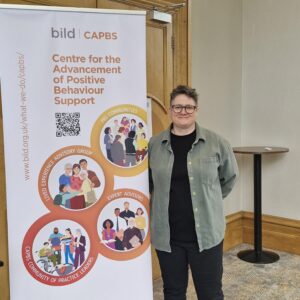 Cairi McKenzie smiling at the camera, standing beside a Bild banner for the Centre for the Advancement of Positive Behaviour Support.