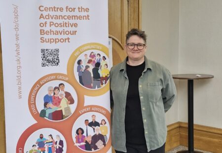 Cairi McKenzie smiling at the camera, standing beside a Bild banner for the Centre for the Advancement of Positive Behaviour Support.