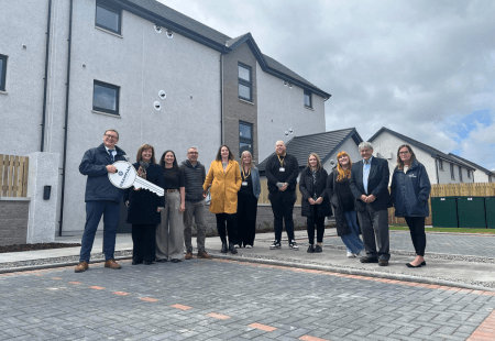 A group of people stand on a paved area outside newly built apartment buildings, posing for a photo, with one person holding a large ceremonial key.