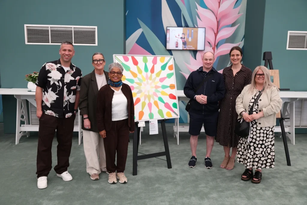 Six people stand together in an indoor gallery space beside a brightly coloured abstract painting displayed on an easel, with additional artworks and a screen visible in the background.