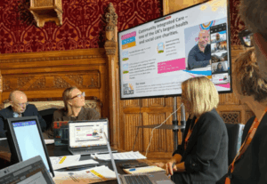 A roundtable meeting in a wood-panelled Westminster room, with participants seated around a table using laptops while viewing a large screen displaying a presentation about Community Integrated Care. Edd Terrey is also on screen as a video call participant.