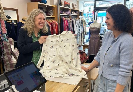 Gillian and a person we support are stood together at the counter in our Inspire…By shop. They are both smiling at each other and holding up a patterned shirt, with racks of second-hand clothes behind them and a till in the foreground.