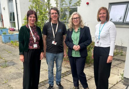 Gillian stands with three of our colleagues, all smiling to camera for a group photo. They’re together outdoors in a courtyard outside a building, with plants, windows and paving visible around them.
