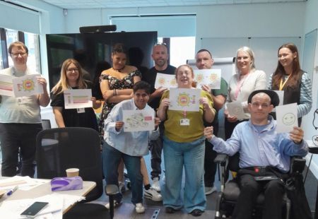 A team of our Quality Advisors stand in an open office space all smiling to camera and holding up various posters with different signs on them.