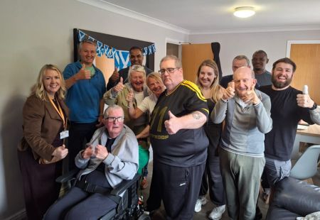 A group of 12 colleagues and people we support stand in a communal room all smiling and thumbs up to camera. There's bunting of Scottish flags in the background.