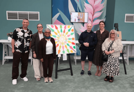 Six people stand together in an indoor gallery space beside a brightly coloured abstract painting displayed on an easel, with additional artworks and a screen visible in the background.