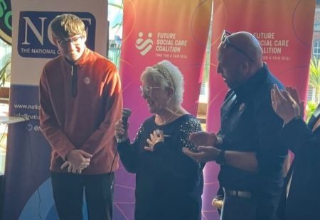 Two people we support and a colleague stood together, in front of orange event banners. Susan is in the centre and speaking into the microphone, whilst Matt and Edd stand either side.