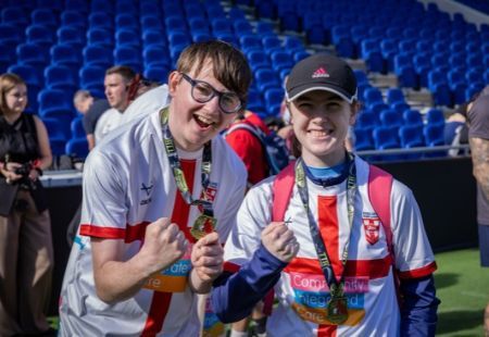 Matt Price and Katie Richardson are smiling to camera and cheering. They are on a football pitch with blue seats behind them.