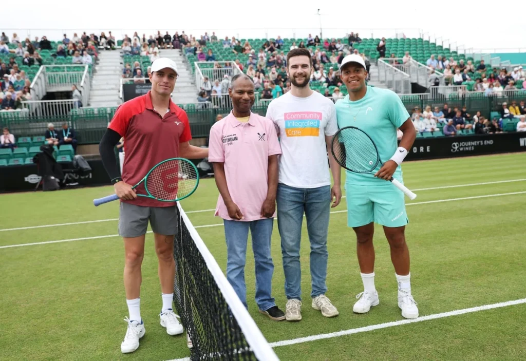 Two tennis players and two representatives from Community Integrated Care pose together on a grass court before a match, with spectators watching from the stands behind them.