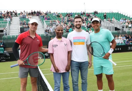 Two tennis players and two representatives from Community Integrated Care pose together on a grass court before a match, with spectators watching from the stands behind them.