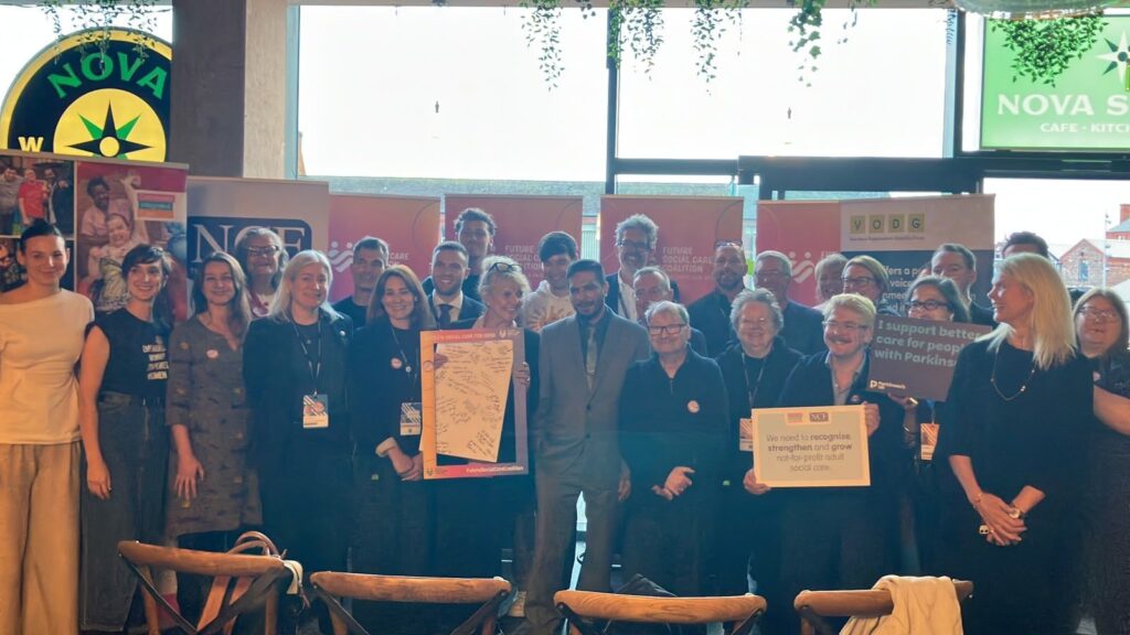 A large group of people stand together in a cafe cheering. They are holding signs campaigning for social care.